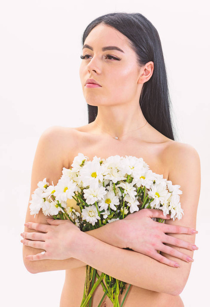 Woman with smooth healthy skin looks attractive. Lady covers breasts with flowers, isolated on white. Girl on calm face naked holds chamomile flowers in front of breasts. Natural cosmetics concept - Foto, Bild