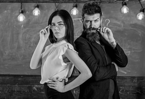 Man with beard and teacher in eyeglasses stand back to back, chalkboard on background. Lady and hipster working together in school. Teacher and schoolmaster look confident. Teamwork concept - Foto, afbeelding