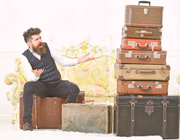 Macho elegant on surprised face sits shocked near pile of vintage suitcase. Luggage and travelling concept. Man, butler with beard and mustache delivers luggage, luxury white interior background - Foto, afbeelding
