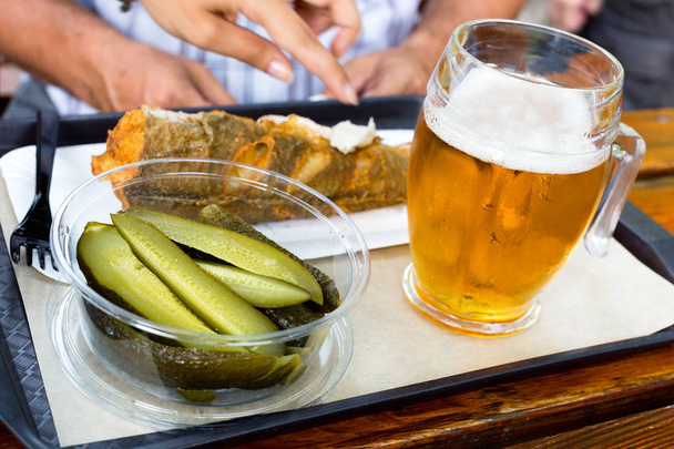 Νόστιμα τηγανητά ψάρια, τουρσί αγγούρια και παγωμένο ποτήρι βαρελίσια μπίρα σε ένα δίσκο σε ξύλινο τραπέζι στην εξωτερική καφετέρια - Φωτογραφία, εικόνα