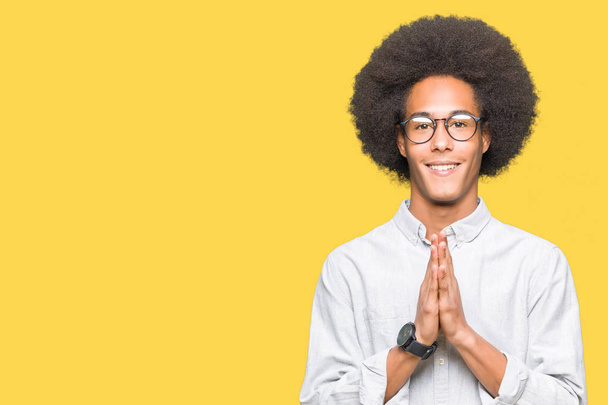 Jovem afro-americano com cabelo afro usando óculos orando com as mãos juntas pedindo perdão sorrindo confiante
. - Foto, Imagem