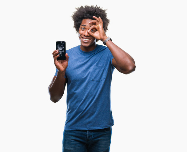 Афро-американец держит сломанный смартфон на изолированном фоне со счастливым лицом улыбаясь делает хорошо знак с рукой на глазу глядя сквозь пальцы
 - Фото, изображение
