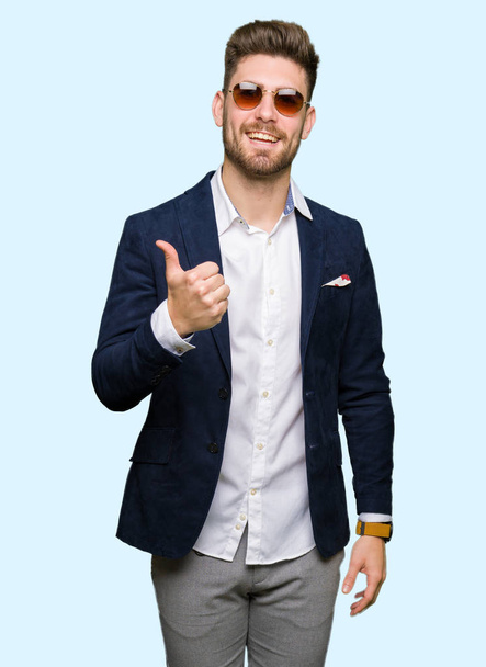 Young handsome elegant man wearing sunglasses and fashion blazer doing happy thumbs up gesture with hand. Approving expression looking at the camera with showing success. - Photo, Image