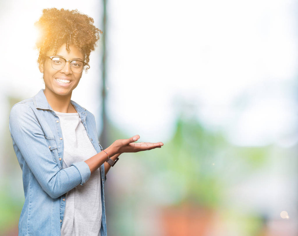 Mooie jonge african american vrouw het dragen van bril geïsoleerd achtergrond wijst naar de kant met de hand en open palm, presenteren ad glimlachend gelukkig en vol vertrouwen - Foto, afbeelding