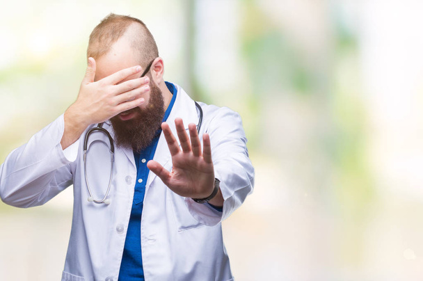 Young caucasian doctor man wearing medical white coat over isolated background covering eyes with hands and doing stop gesture with sad and fear expression. Embarrassed and negative concept. - Photo, Image