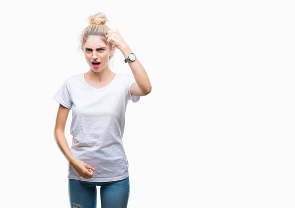 Young beautiful blonde woman wearing white t-shirt over isolated background angry and mad raising fist frustrated and furious while shouting with anger. Rage and aggressive concept. - Photo, Image