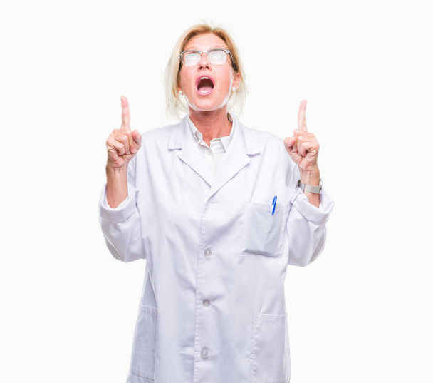 Middle age blonde therapist woman wearing white coat over isolated background amazed and surprised looking up and pointing with fingers and raised arms. - Foto, immagini