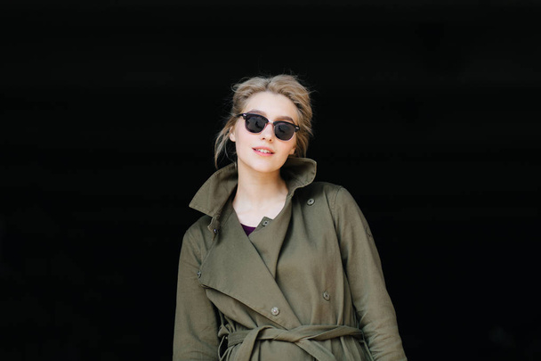 portrait of young blonde woman in stylish coat and sunglasses on black background - Фото, изображение