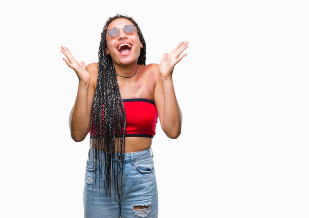 Young gevlochten haar Afrikaanse Amerikaan met geboorte merk zonnebril dragen over geïsoleerde achtergrond vieren gek en verbaasd voor succes met armen verhoogd en open ogen schreeuwen opgewonden. Winnaar concept - Foto, afbeelding