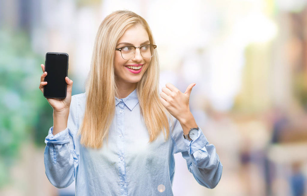 Jovem mulher de negócios loira bonita mostrando tela do smartphone sobre fundo isolado apontando e mostrando com o polegar até o lado com o rosto feliz sorrindo
 - Foto, Imagem