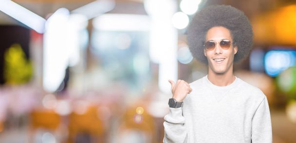 junger afrikanisch-amerikanischer Mann mit Afro-Haaren trägt Sonnenbrille, lächelt mit glücklichem Gesicht und zeigt mit erhobenem Daumen zur Seite. - Foto, Bild