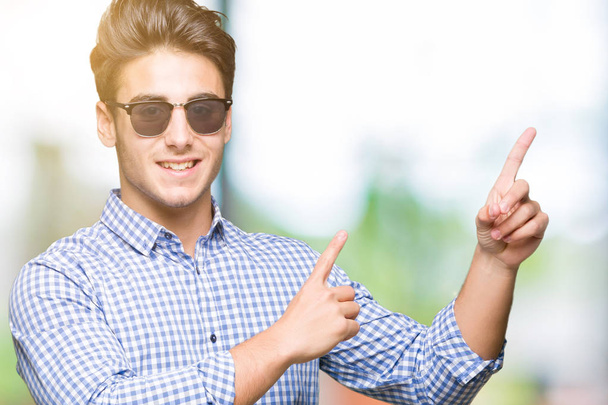 Mladý pohledný muž nosí sluneční brýle izolované pozadí s úsměvem a při pohledu na fotoaparát s oběma rukama a prsty přejdete na stranu. - Fotografie, Obrázek