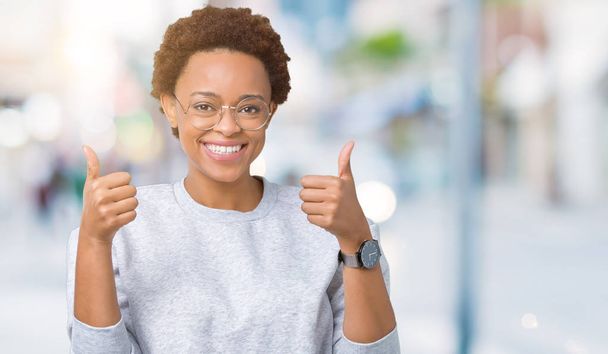 Young beautiful african american woman wearing glasses over isolated background success sign doing positive gesture with hand, thumbs up smiling and happy. Looking at the camera with cheerful expression, winner gesture. - Photo, Image