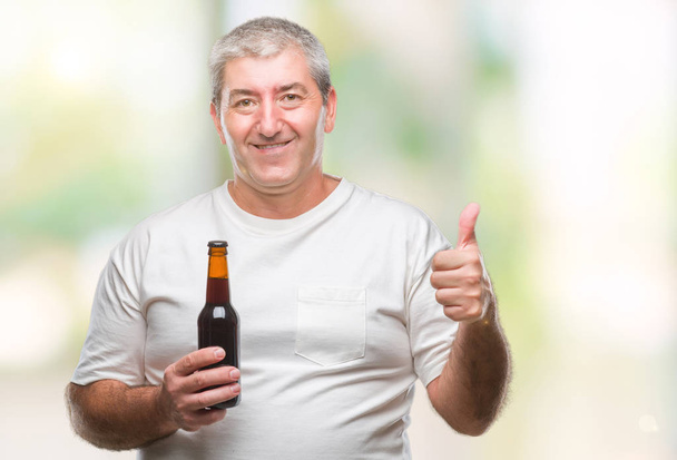 Przystojny mężczyzna senior picia butelka piwa na białym tle szczęśliwy z uśmiechem robi znak ok, kciuk palcami, bardzo dobry znak - Zdjęcie, obraz