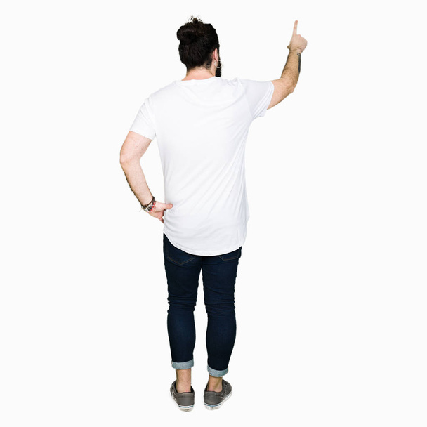 junger Hipster-Mann mit langen Haaren und Bart in lässigem weißem T-Shirt, der rückwärts mit der Fingerhand posiert - Foto, Bild