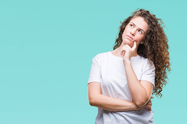 Beautiful brunette curly hair young girl wearing casual t-shirt over isolated background with hand on chin thinking about question, pensive expression. Smiling with thoughtful face. Doubt concept. - Foto, Imagem