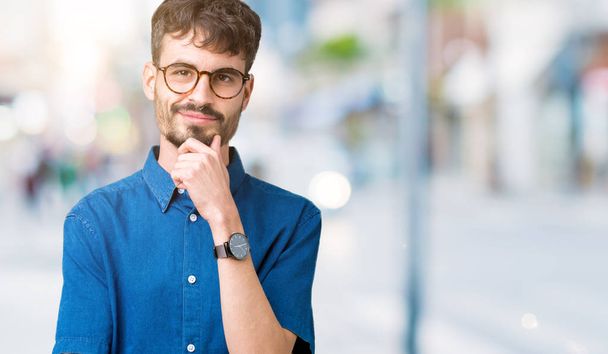 Young handsome man wearing glasses over isolated background looking confident at the camera with smile with crossed arms and hand raised on chin. Thinking positive. - Photo, Image