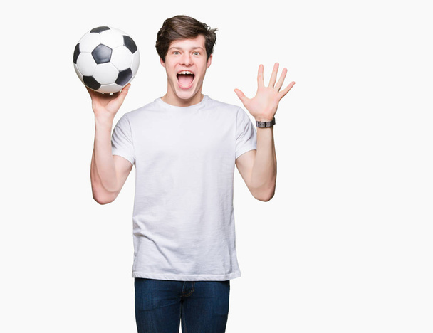 Młody człowiek gospodarstwa piłka nożna Piłka nożna na na białym tle bardzo szczęśliwy i podekscytowany, zwycięzca wyrażenie świętuje zwycięstwo krzyczy z uśmiechem i ręce - Zdjęcie, obraz