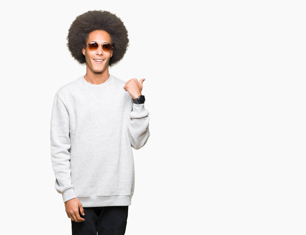 Jonge Afro-Amerikaanse man met afro haar dragen van een zonnebril glimlachend met blij gezicht kijken en naar de kant met duim omhoog. - Foto, afbeelding