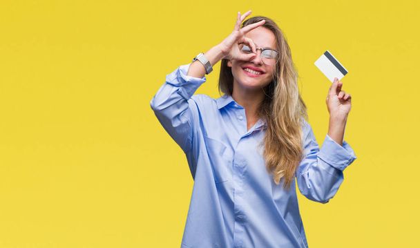 Młody piękny blond kobieta trzymając kartę kredytową na białym tle z szczęśliwy twarz uśmiechający się robi ok znak ręką na oko patrząc przez palce - Zdjęcie, obraz