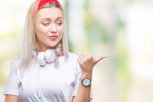 Молода блондинка в навушниках слухає музику над ізольованим фоном, посміхаючись із щасливим обличчям, дивлячись і вказуючи на бік з великим пальцем вгору
. - Фото, зображення