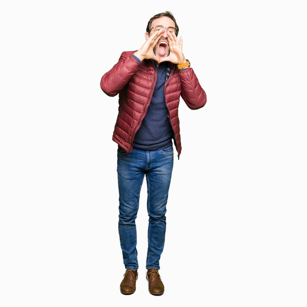 Homem bonito de meia idade vestindo óculos e casaco de inverno gritando irritado em voz alta com as mãos sobre a boca
 - Foto, Imagem