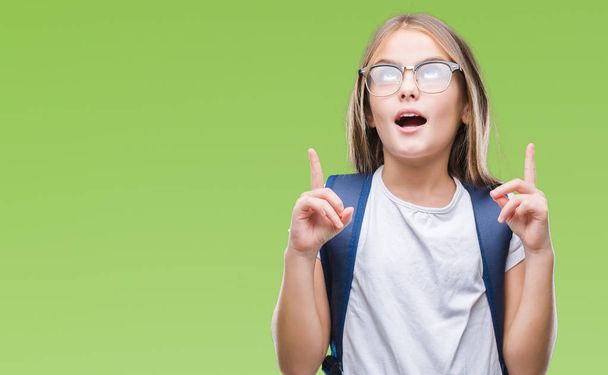 Young beautiful smart student girl wearing backpack over isolated background amazed and surprised looking up and pointing with fingers and raised arms. - 写真・画像