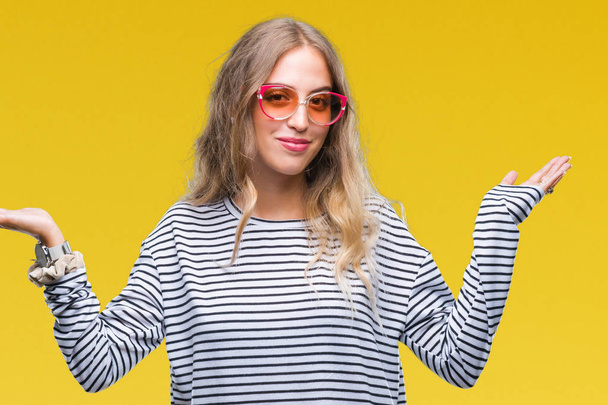 Beautiful young blonde woman wearing sunglasses over isolated background clueless and confused expression with arms and hands raised. Doubt concept. - Zdjęcie, obraz