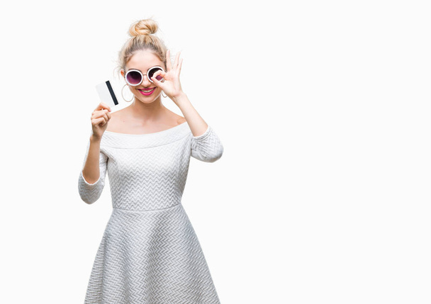 Jonge mooie blonde vrouw die houdt van creditcard over geïsoleerde achtergrond met blij gezicht lachend doen ok teken met de hand op oog kijken door de vingers - Foto, afbeelding
