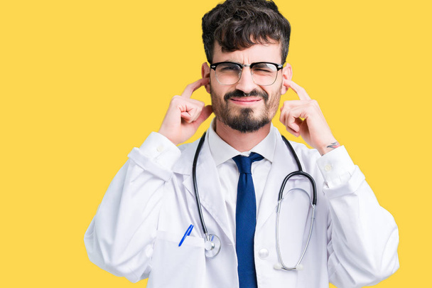 Young doctor man wearing hospital coat over isolated background covering ears with fingers with annoyed expression for the noise of loud music. Deaf concept. - Photo, Image