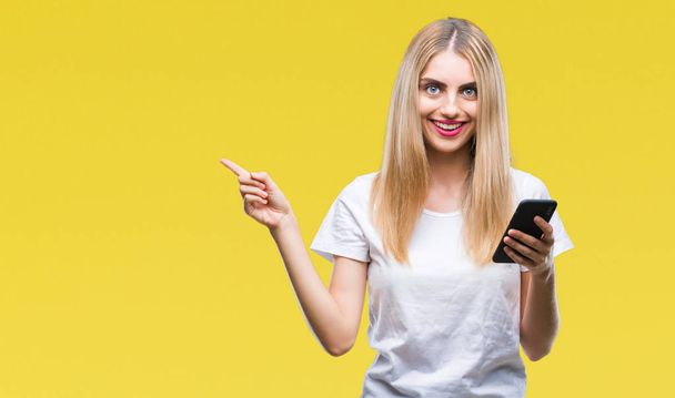 Jonge mooie blonde vrouw met smartphone via geïsoleerde achtergrond erg blij met de hand en de vinger te wijzen naar de kant - Foto, afbeelding