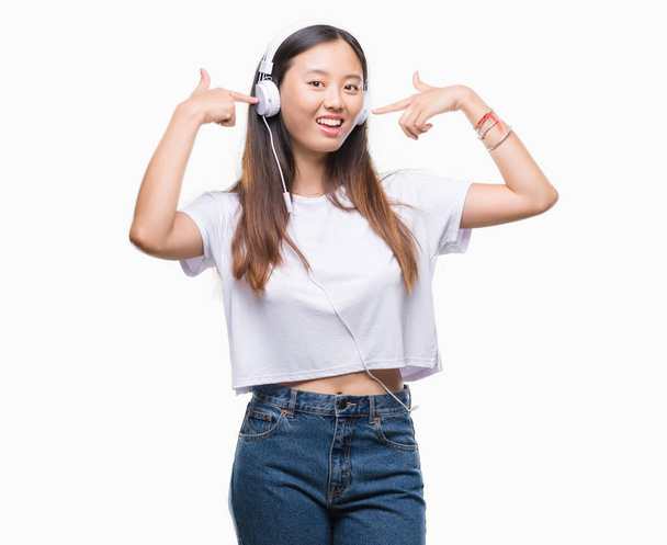 Молода азіатська жінка слухає музику в навушниках ізольований фон дуже щасливий, вказуючи рукою і пальцем
 - Фото, зображення