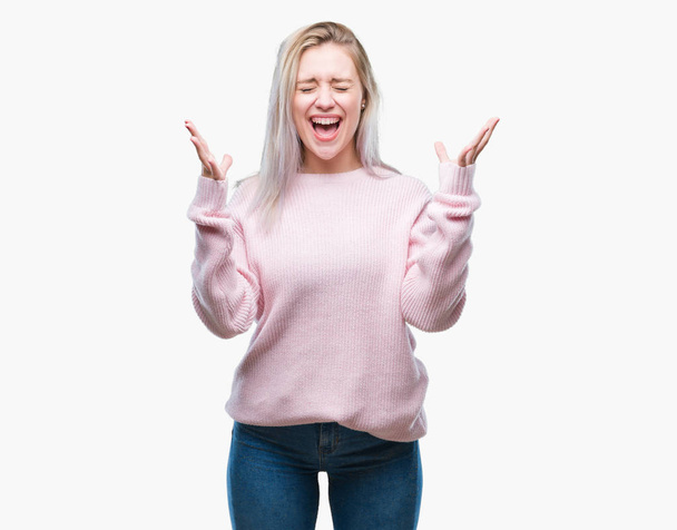 Νεαρή ξανθιά γυναίκα, φορώντας χειμώνα πουλόβερ πάνω από απομονωμένες φόντο γιορτάζει τρελό και κατάπληκτος για την επιτυχία με τα χέρια υψωμένα και ανοίξτε τα μάτια ουρλιάζοντας ενθουσιασμένος. Νικητής έννοια - Φωτογραφία, εικόνα