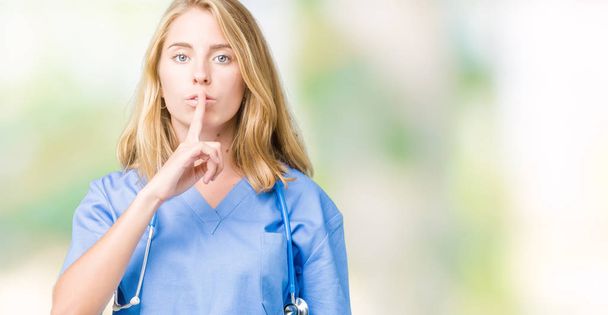 Όμορφη νεαρός γιατρός γυναίκα που φοράει ιατρική στολή πέρα από το απομονωμένο υπόβαθρο ζητώντας να είναι ήσυχη με το δάχτυλο στα χείλη. Σιωπή και μυστικό έννοια. - Φωτογραφία, εικόνα