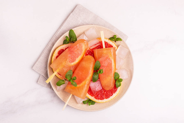 Homemade grapefruit popsicle with ripe grapefruit slices and fresh mint on light marble background. Healthy desserts. Lactose free ice cream. - Фото, изображение