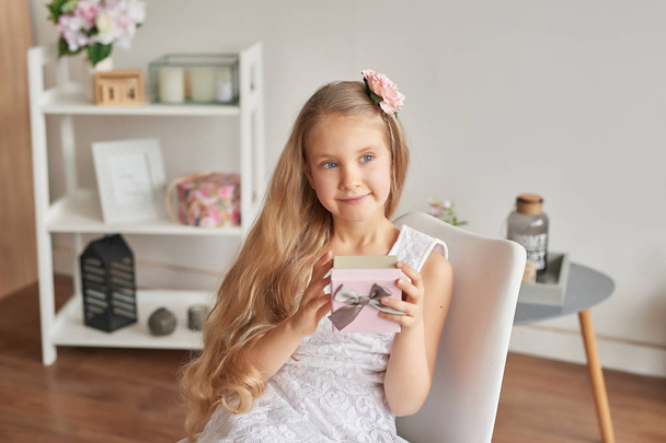 menina em um vestido branco senta-se em uma cadeira e mantém um presente para a mãe
 - Foto, Imagem