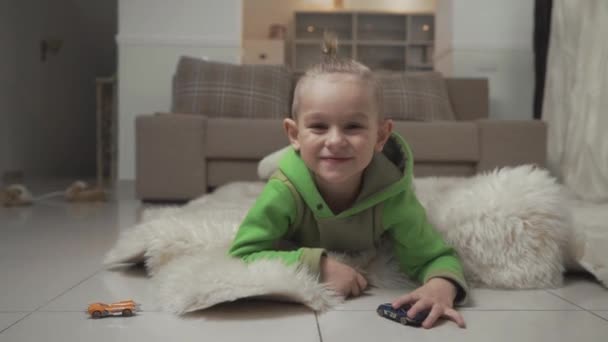 Portrait of little smilling boy with stylish haircut playing with toycars lying on the floor on fluffy carpet. - Felvétel, videó