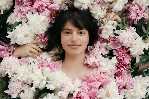 Portrait of boho woman with natural makeup lying in peonies. Creative floral photo. Aroma and spa concept. International Womens Day. Beautiful brunette girl in many pink and white flowers - Foto, afbeelding