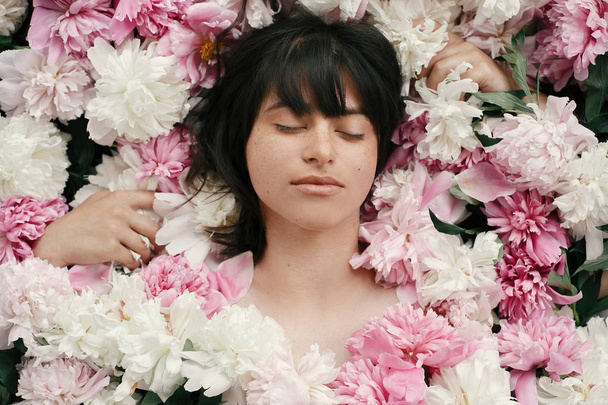 Portrait of boho woman with natural makeup lying in peonies. Creative floral photo. Aroma and spa concept. International Womens Day. Beautiful brunette girl in many pink and white flowers - Zdjęcie, obraz