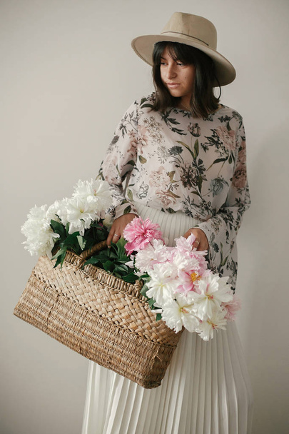 Sinnliches Porträt eines Boho-Mädchens mit rosa und weißen Pfingstrosen in rustikalem Korb. stylische Hipster-Frau mit Hut und Bohème-Blumenkleid, die mit Blumen posiert. Internationaler Frauentag. - Foto, Bild