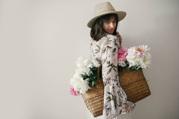 Chica boho en sombrero sosteniendo peonías rosas y blancas en canasta rústica. Mujer hipster con estilo en vestido floral bohemio posando con flores. Día Internacional de la Mujer. Vida en el campo
 - Foto, imagen