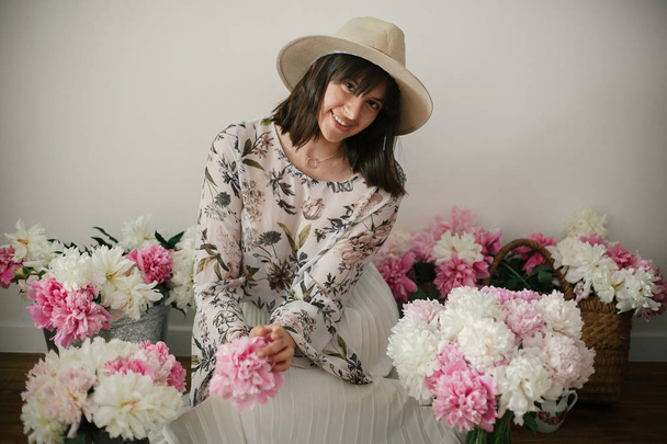 Happy boho girl smiling at pink and white peonies on rustic wooden floor. Stylish hipster woman in hat and bohemian floral dress  with flowers. International Womens Day. Countryside living - Fotografie, Obrázek