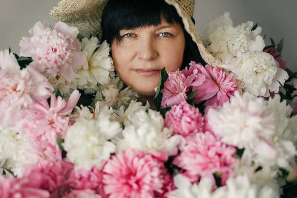 Piękna kobieta starszy w kapelusz pozowanie z wielu kwiaty piwonii. Szczęśliwą matką z wielkim bukietem różowe piwonie od dzieci. Szczęśliwy dzień matki koncepcji. Międzynarodowy Dzień kobiet - Zdjęcie, obraz