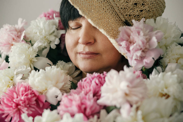 Beautiful elder woman in hat posing with many peony flowers. Happy mother with big bouquet of pink peonies from children. Happy Mothers day concept. International Womens Day - Photo, Image