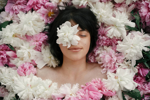 Beautiful brunette girl with white peony on eye lying in pink peonies. Aroma and care concept. International Womens Day. Creative art floral photo. Portrait of boho woman in many flowers - Фото, изображение