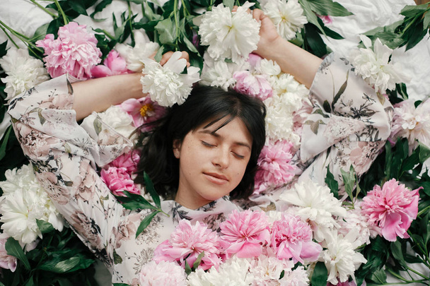 Portrait of boho woman with natural makeup lying in peonies. Creative floral photo. Aroma and spa concept. International Womens Day. Beautiful brunette girl in many pink and white flowers - Fotoğraf, Görsel