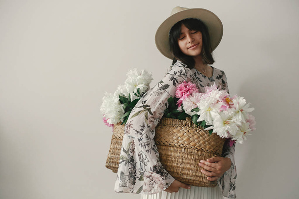 Sensual portrait of boho girl holding pink and white peonies in rustic basket. Stylish hipster woman in hat and bohemian floral dress posing with flowers. International Womens Day. - Photo, image