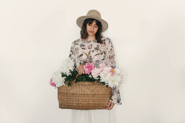 Sensual portrait of boho girl holding pink peonies in rustic basket, isolated on white. Stylish hipster woman in hat and bohemian floral dress posing with flowers. International Womens Day. - Valokuva, kuva