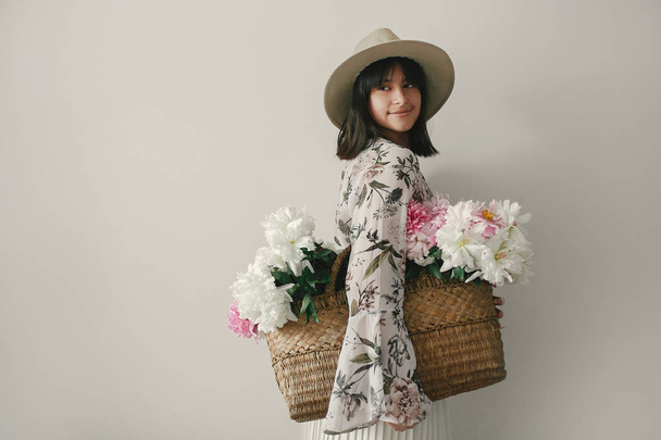 Sensuele portret van boho meisje houdt van roze en witte pioenrozen in rustieke mand. Stijlvolle hipster vrouw in hoed en Boheemse floral dress poseren met bloemen. Internationale dag van de Womens. - Foto, afbeelding