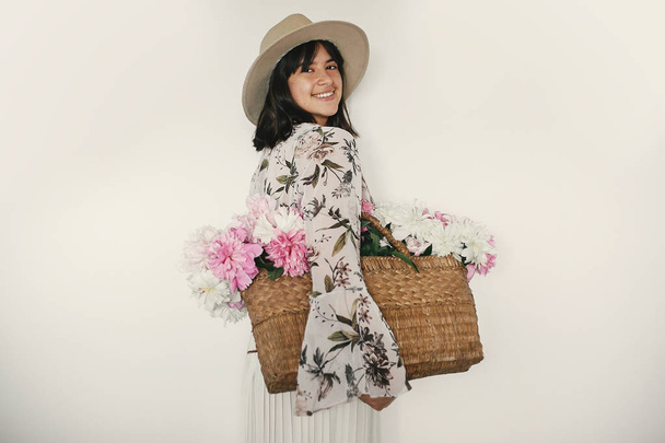 Happy boho girl and holding pink and white peonies in rustic basket. Stylish hipster woman in hat and bohemian floral dress smiling with flowers. International Womens Day. Countryside living - Photo, Image